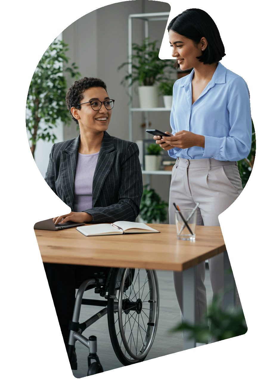 Foto de duas colegas de trabalho em um ambiente de escritório. Uma delas tem cabelos curtos e cacheados, usa óculos e está sentada em sua cadeira de rodas. A outra está em pé ao lado dela, tem cabelo liso chanel escuro. Foto de duas colegas de trabalho em um ambiente de escritório. Uma delas tem cabelos curtos e cacheados, usa óculos e está sentada em sua cadeira de rodas. A outra está em pé ao lado dela, tem cabelo liso chanel escuro.